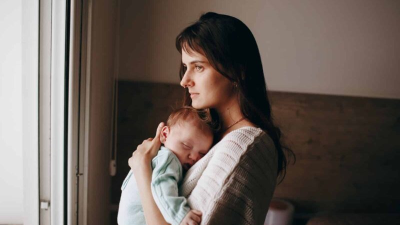 New mother holding her sleeping baby while gazing out a window, a quiet moment that can hide postpartum depression