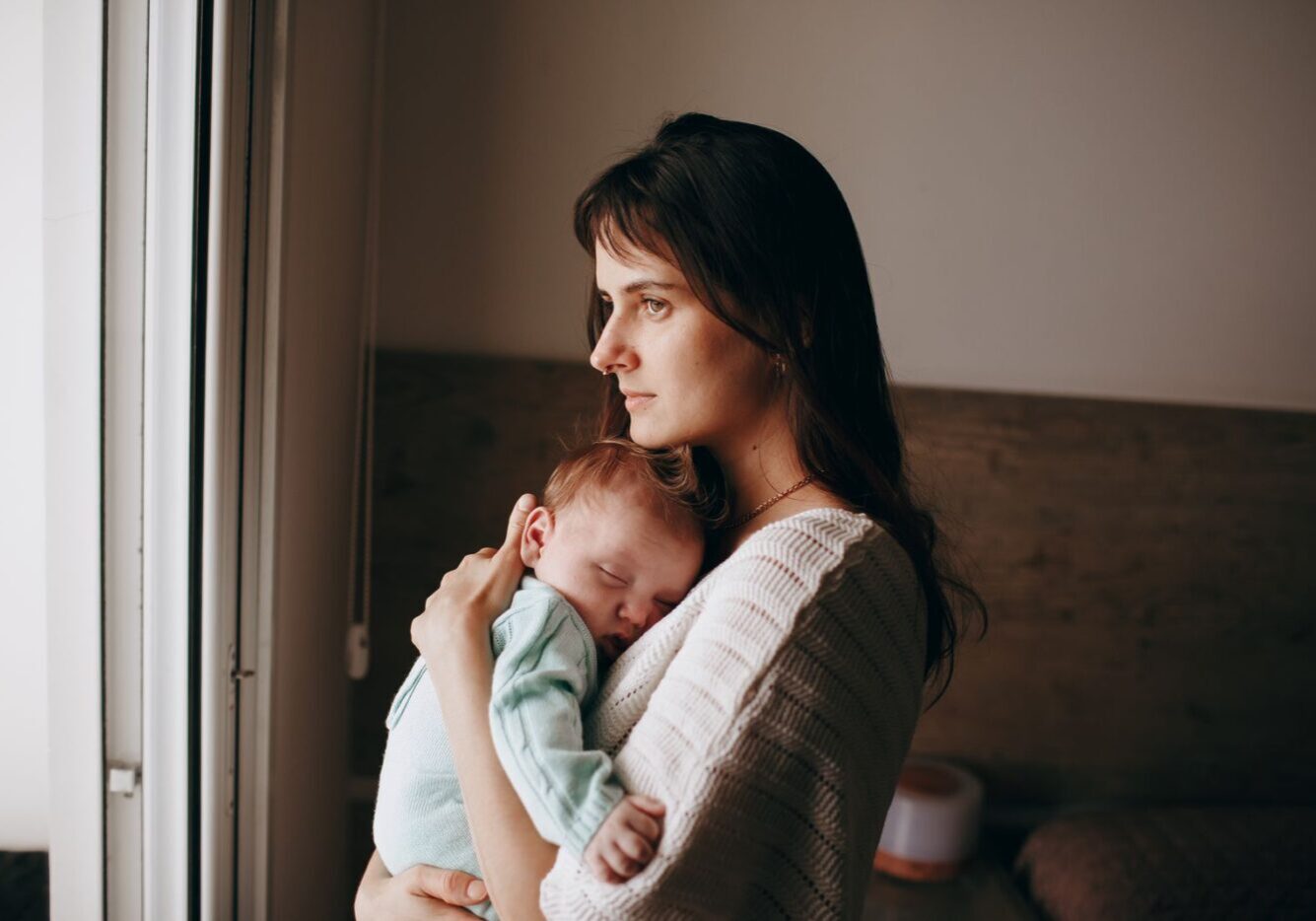 New mother holding her sleeping baby while gazing out a window, a quiet moment that can hide postpartum depression